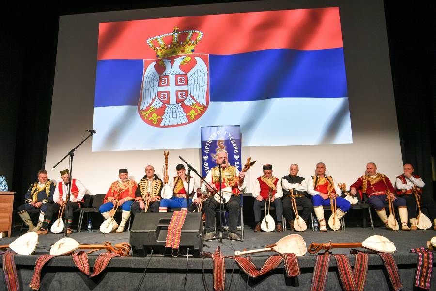 Гусларско вече Гуслари на сцени. Један гусла у првом плану, остали седе иза њега и слушају га. Сцена је украшена појасевима и гуслама. На позадини је застава Републике Србије (црвена, плава и бела са грбом двоглавог орла са круном)