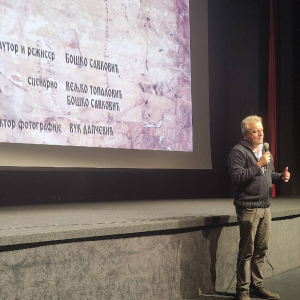 Човек говори испред бине. Иза њега, на платну, је филм „У почетку беше реч”