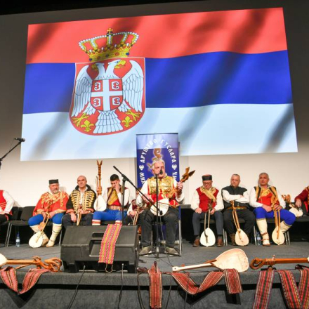 Гуслари на сцени. Један гусла у првом плану, остали седе иза њега и слушају га. Сцена је украшена појасевима и гуслама. На позадини је застава Републике Србије (црвена, плава и бела са грбом двоглавог орла са круном)