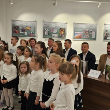 Деца певају у хору, а иза њих седе одрасли који говоре на представљању књиге